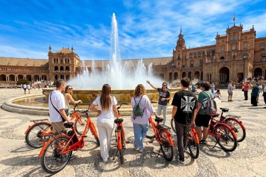 A group of people with bicycles in front of a historic building with a fountain. 1