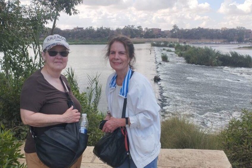 Two people standing by a river with greenery and a distant building in the background. 1