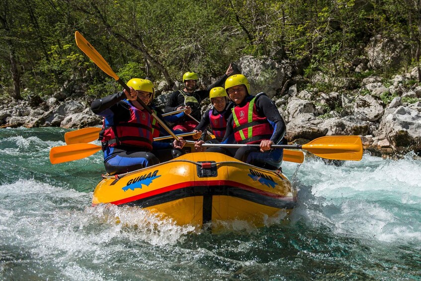 A group of people in a yellow inflatable raft with orange paddles, navigating a river surrounded by rocks and greenery. 1