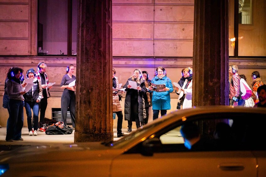 A group of people standing on a sidewalk, some holding papers, with a building in the background. 2