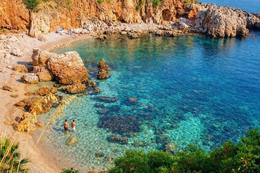 A rocky coastal landscape with clear turquoise water, people walking along the shore, and lush greenery in the foreground. 5