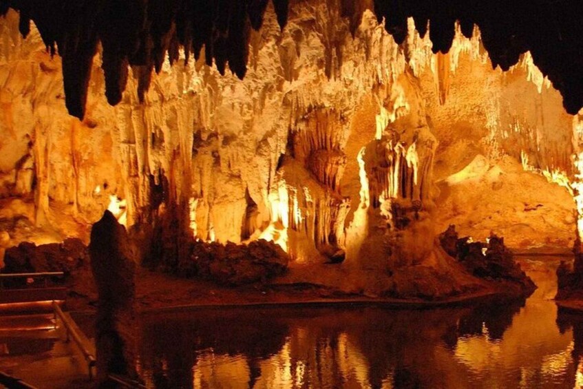 A cave illuminated by artificial lighting, with stalactites and stalagmites, and a reflective water surface in the foreground. 3