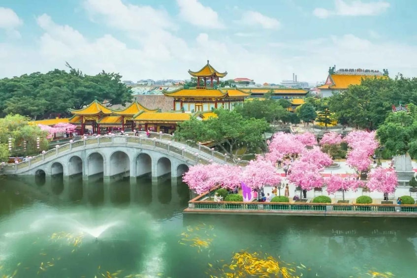 A traditional Chinese garden with a bridge over water, cherry blossoms, and pagoda-style buildings. 1