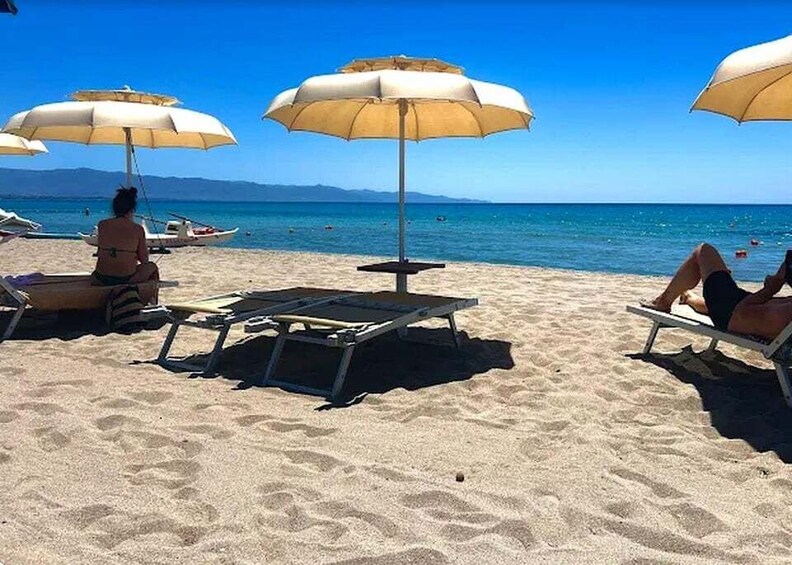 A beach with sun loungers and umbrellas, calm sea, and mountains in the distance. 2