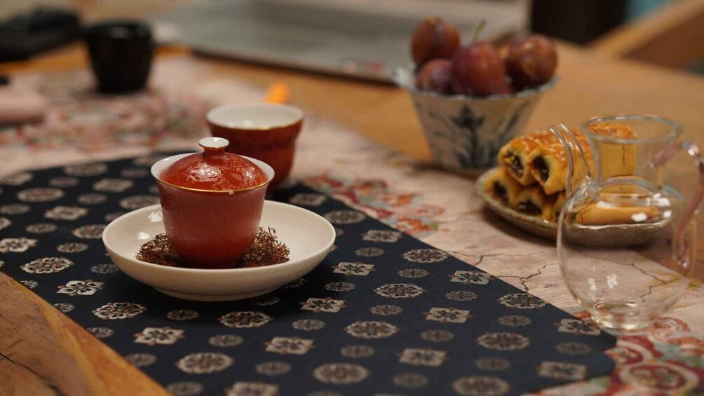 A table with a patterned tablecloth, a bowl of fruit, a glass of a clear liquid, and a cup with a lid. 2
