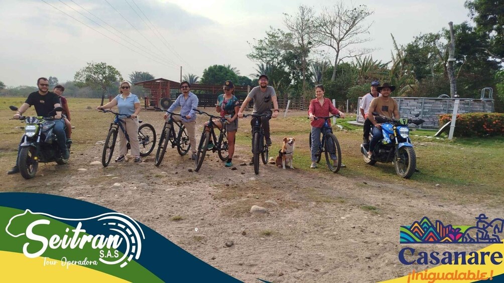 A group of people on bicycles and motorcycles in an outdoor setting with a dirt ground, trees, and a building in the background. 3