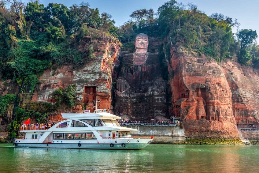 A large rock face with a carved Buddha statue, a boat with passengers, and a forested cliff. 1