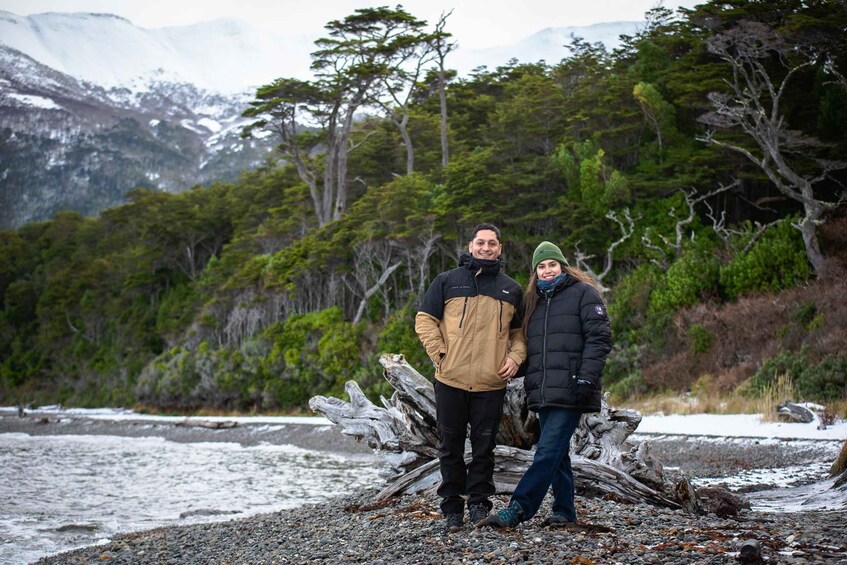 Two people standing on a rocky shore with a forest and mountains in the background. 2