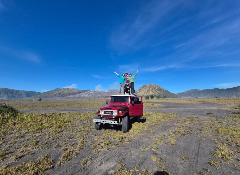 A red truck parked on a dirt road with a group of people standing on top, surrounded by a flat landscape and distant mountains under a clear blue sky. 2