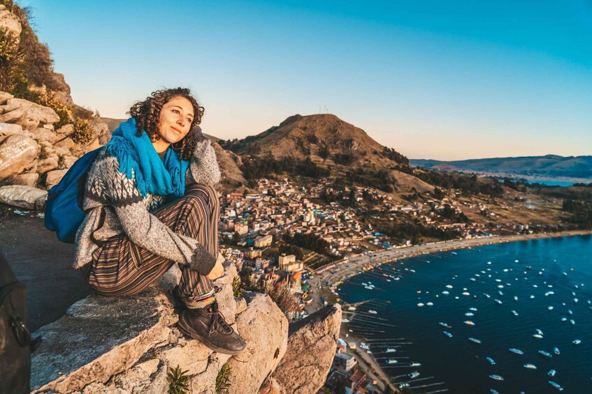A person sitting on a rocky ledge overlooking a coastal town with a clear view of the sea and boats. 1