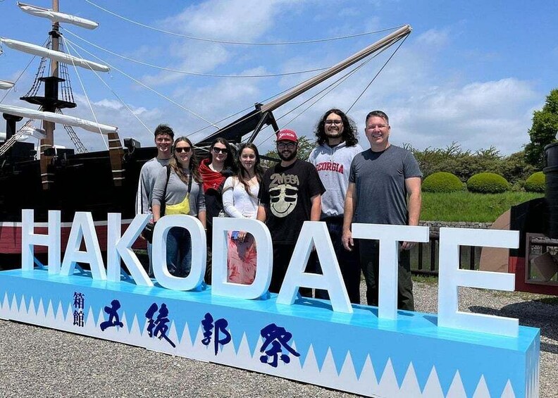 A group of people standing in front of a sign that reads 'HAKODATE' with Japanese characters below. 1