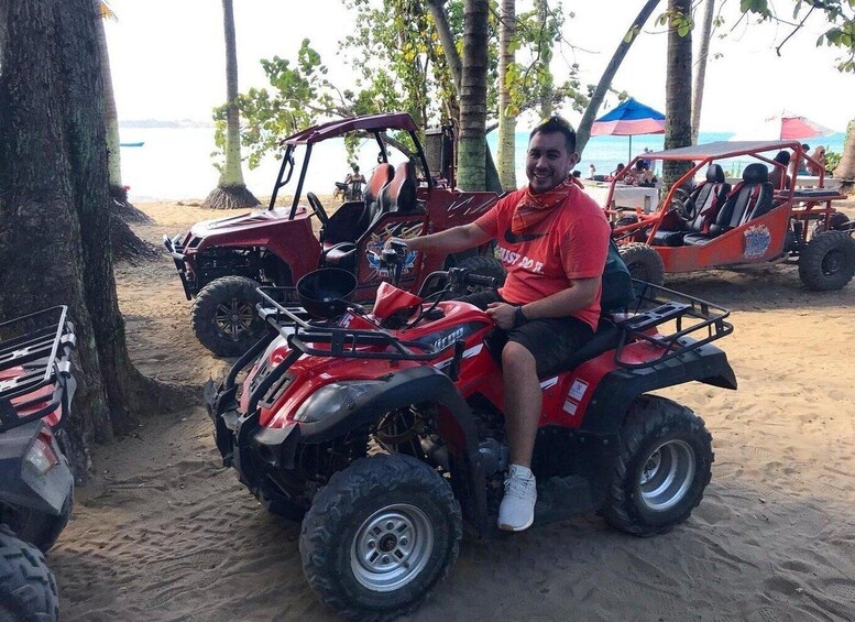 A person riding a red ATV on a sandy beach with palm trees and a blue umbrella in the background. 1