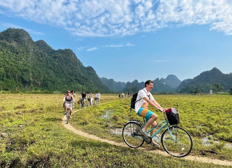 A group of people cycling on a dirt path through a grassy field with mountains in the background. 1