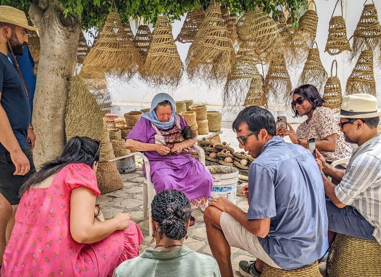 A group of people gathered around a woman seated on a chair, with baskets and woven items displayed in the background. 2