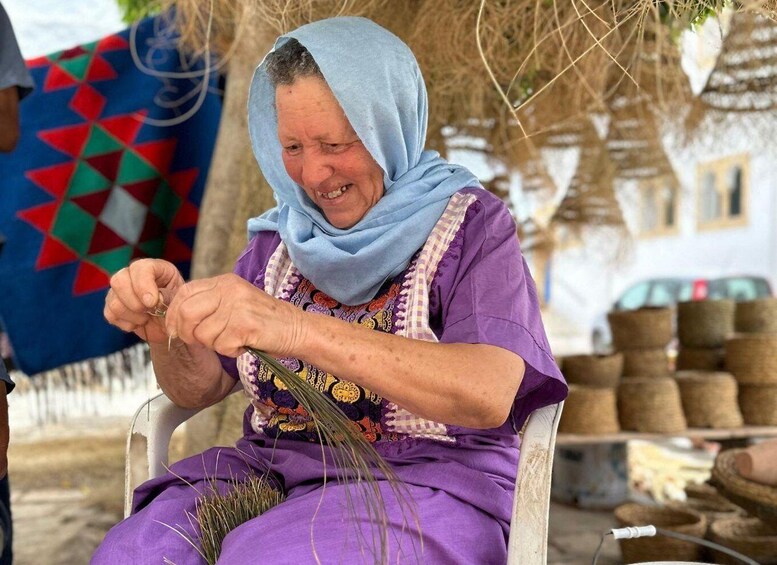 An elderly woman seated, engaged in needlework, with a patterned garment and a blue headscarf. 5