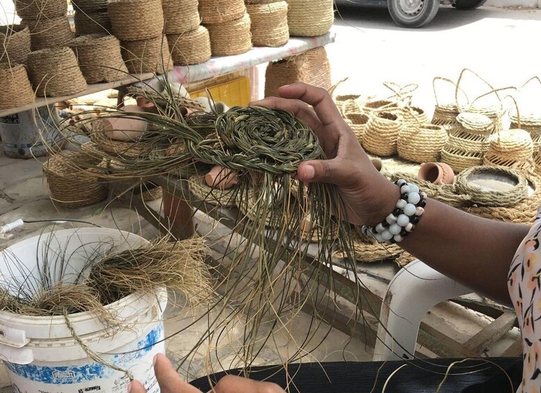 A person holding a bundle of dried grass, with woven baskets and a bucket in the foreground. 5