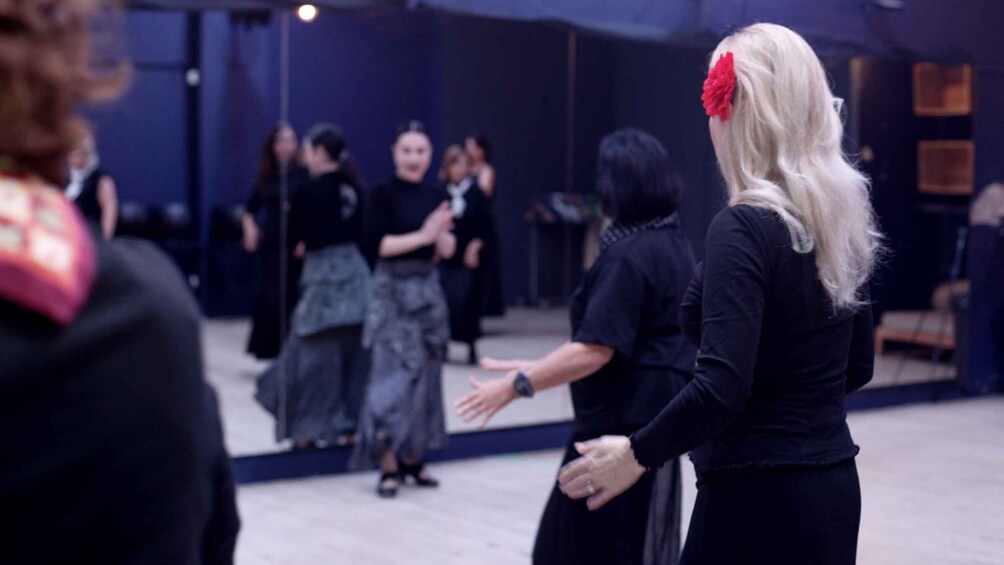 A group of people in a dimly lit indoor setting, possibly a dance studio or event space, with a reflective floor and a dark blue wall in the background. 1