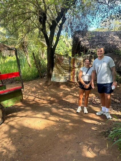 Two people standing on a dirt path with trees and a bench in a park. 2