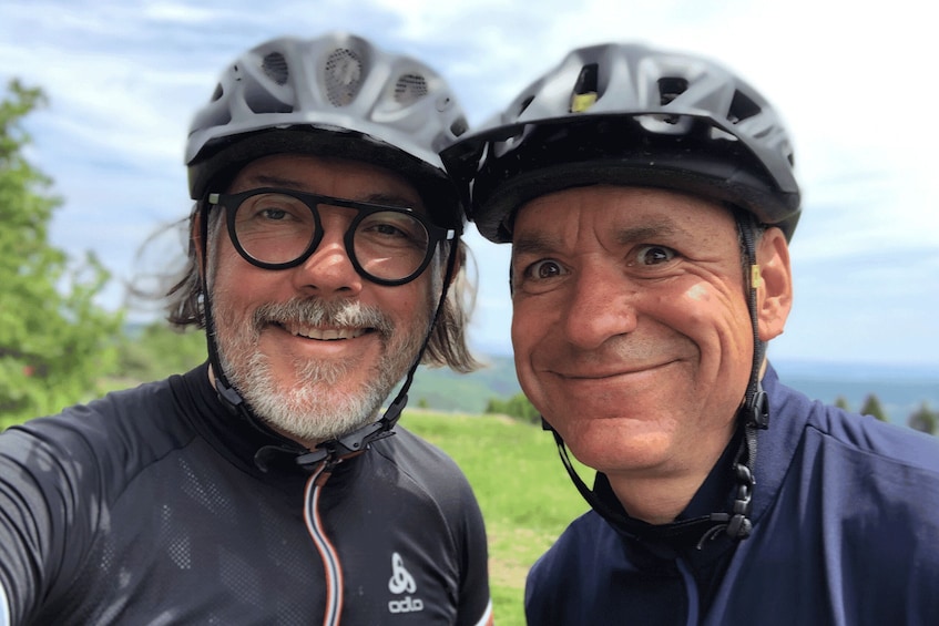 Two people wearing helmets and cycling attire, standing outdoors with a grassy field and a body of water in the background. 1