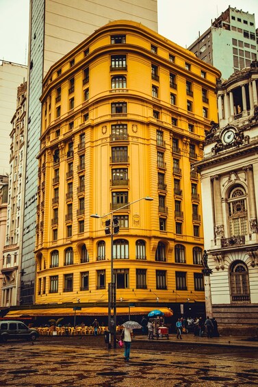 A yellow building with multiple windows and balconies, a clock tower, and a street with people and umbrellas. 2