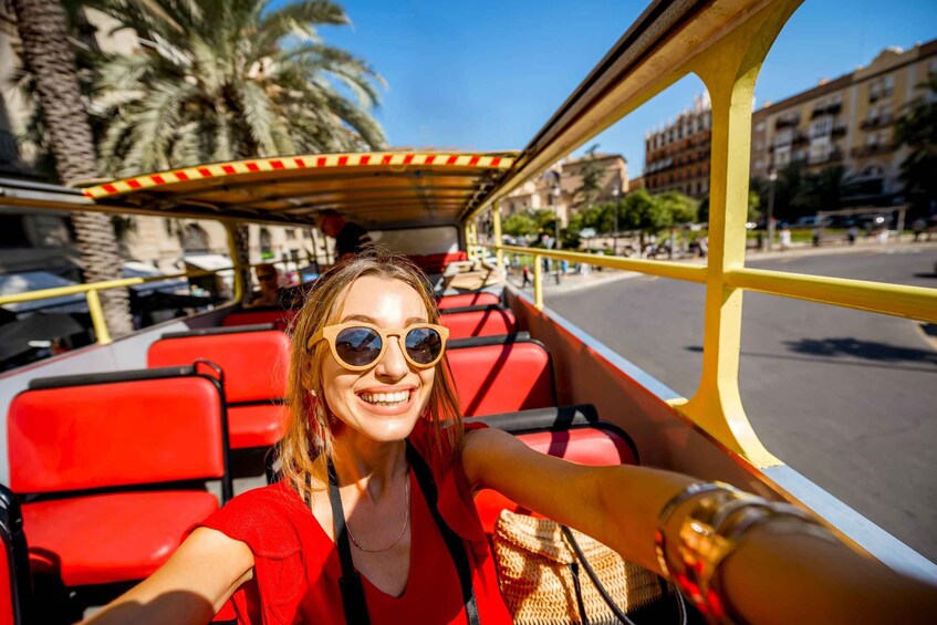 A person seated on a red bus with a yellow roof, palm trees, and buildings in the background. 1