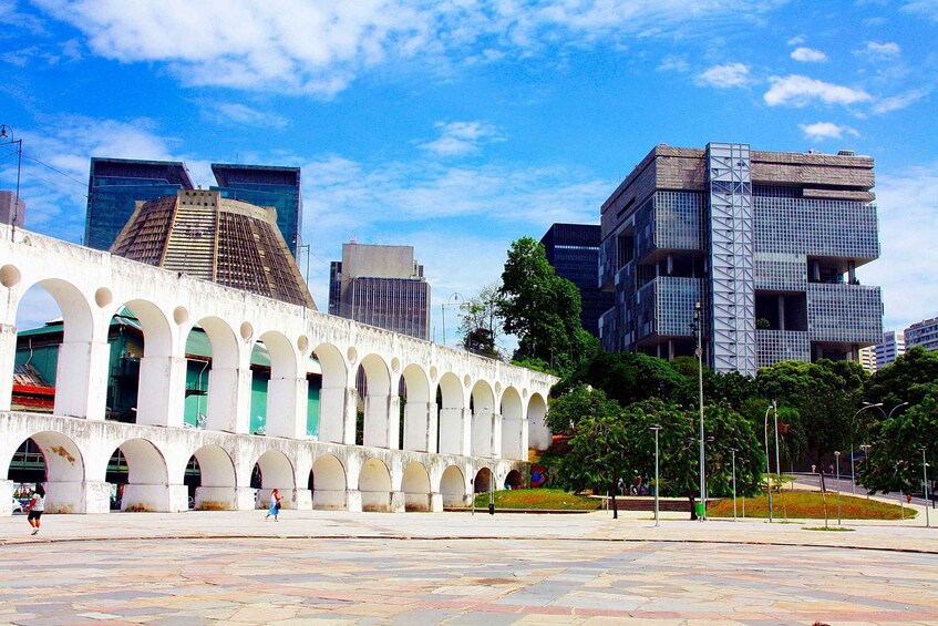 A plaza with a row of arches, modern buildings, and a clear blue sky. 4