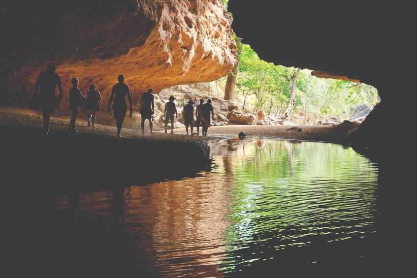 A group of people walking along a riverbank near a cave. 5