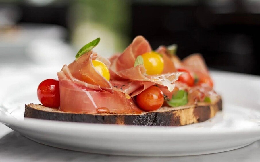 A plate with a slice of toasted bread topped with prosciutto, cherry tomatoes, and a basil leaf. 1