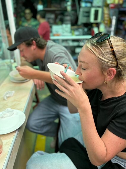 A person sitting at a table with a bowl and spoon, another person in the background, and a store interior with shelves. 2