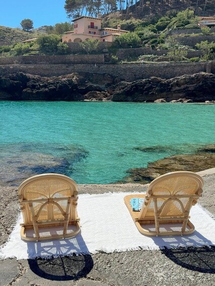 Two wicker chairs on a white mat by a turquoise sea with rocky cliffs in the background. 5