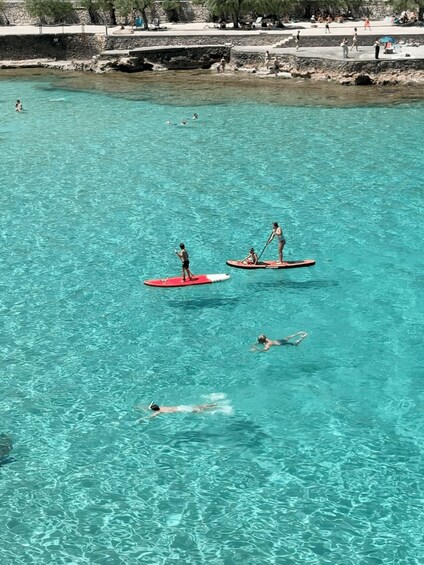 People paddleboarding on a clear turquoise sea with rocky shorelines and a stone wall in the background. 2