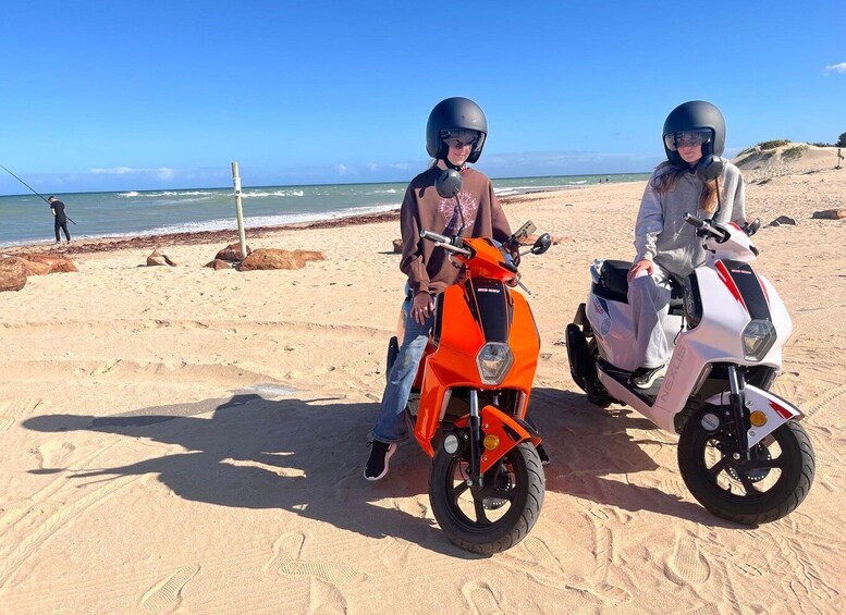 Two people on scooters on a sandy beach with the ocean in the background. 3