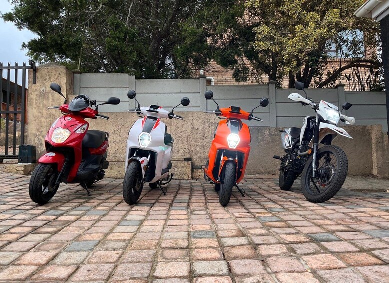 Three scooters parked on a brick pavement with a metal fence and trees in the background. 4