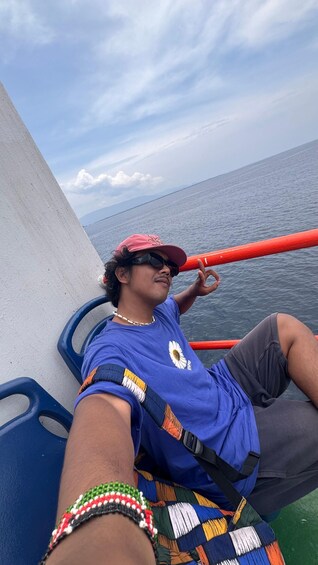 A person reclining on a boat, wearing a blue shirt with a white flower emblem, a red cap, and a colorful beaded bracelet. 3