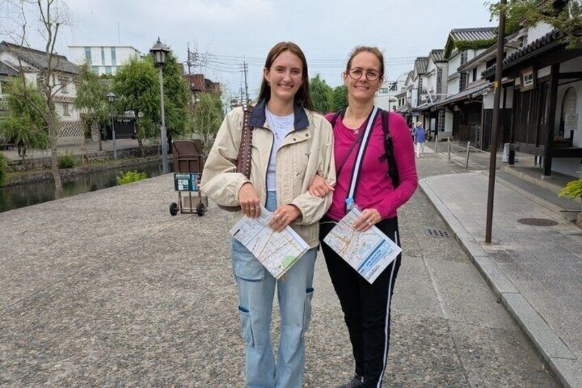 Two people holding maps, standing on a cobblestone path with traditional buildings and a canal in the background. 5
