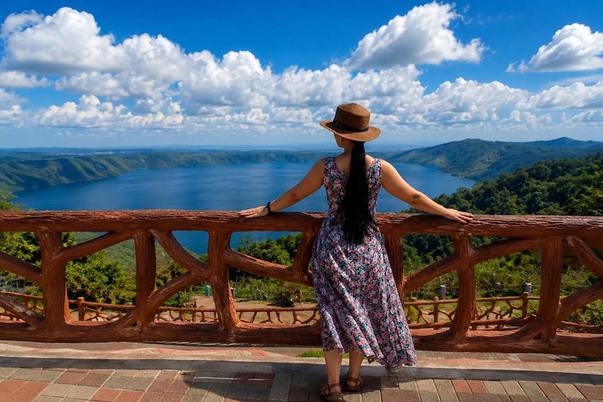 A woman in a floral dress and hat standing on a bridge with a scenic view of a lake and mountains. 5