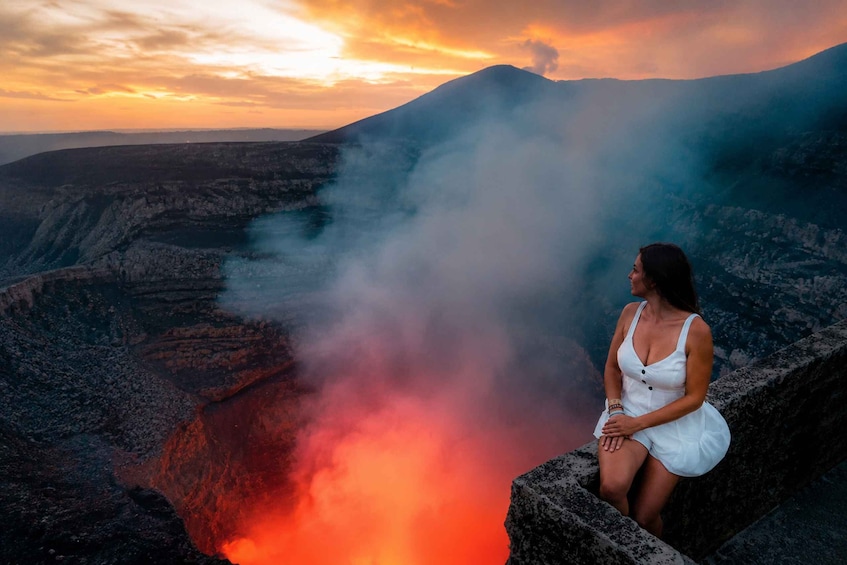 A person sitting on a rock overlooking a volcanic crater with smoke and lava. 2