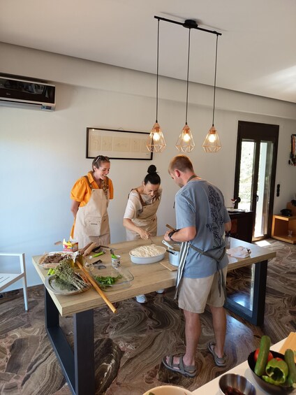 Three people in a kitchen preparing food, with a marble floor, pendant lights, and a framed picture on the wall. 2
