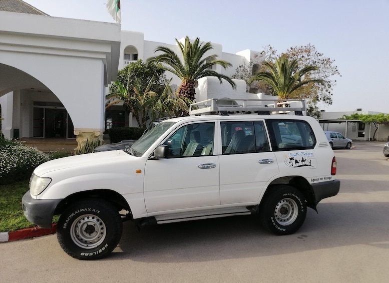 A white SUV with 'Youth Explorer' and 'Agence de Voyage' logos parked in front of a building with arches and palm trees. 1