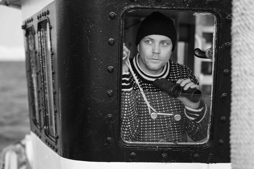 A person in a patterned sweater holding binoculars, looking out from a window with riveted metal. 3