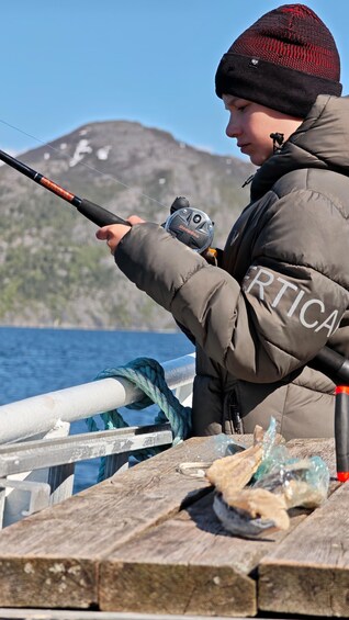 A person fishing with a rod, wearing a beanie and a jacket, by a body of water with a mountainous backdrop. 4