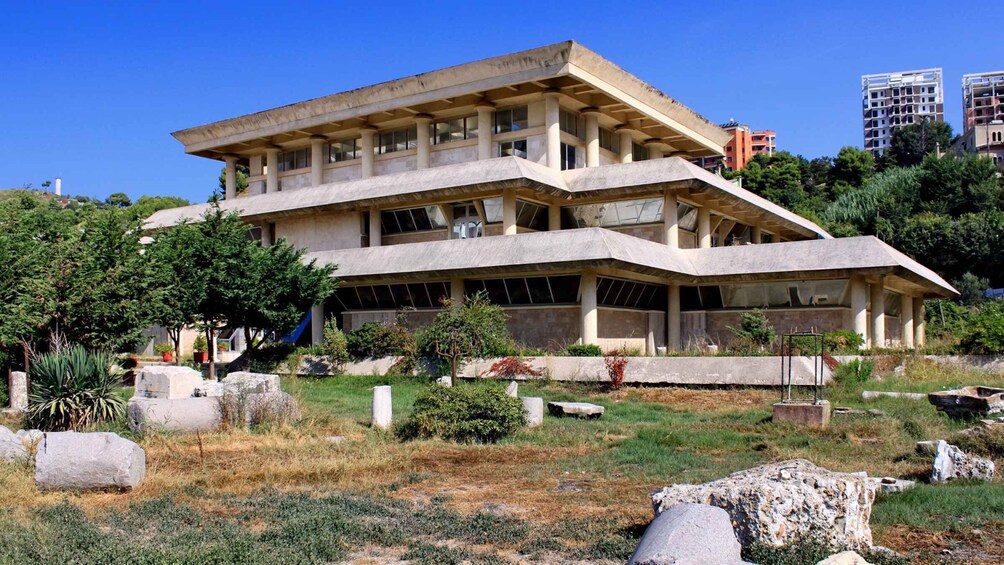 A multi-story concrete building with a sloped roof, surrounded by greenery and rocks. 1