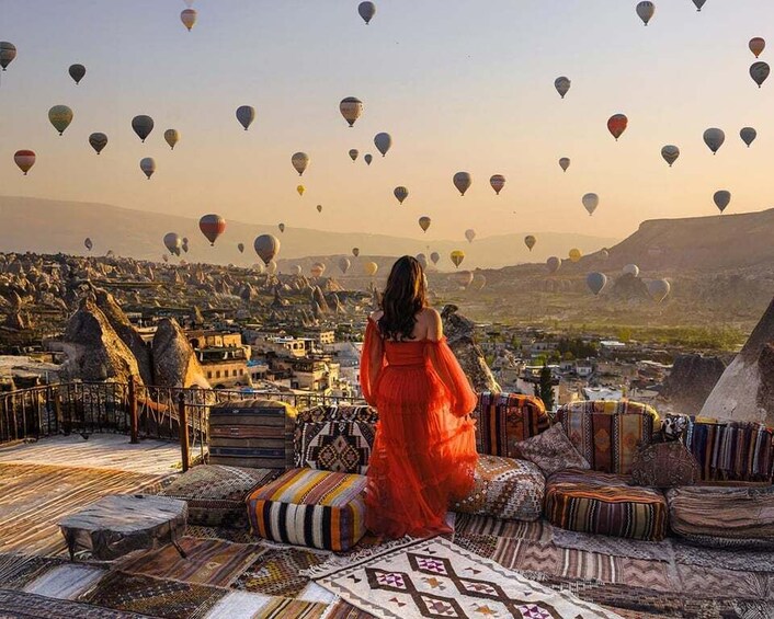 A person in a red dress on a rooftop overlooking a village with hot air balloons in the sky. 3