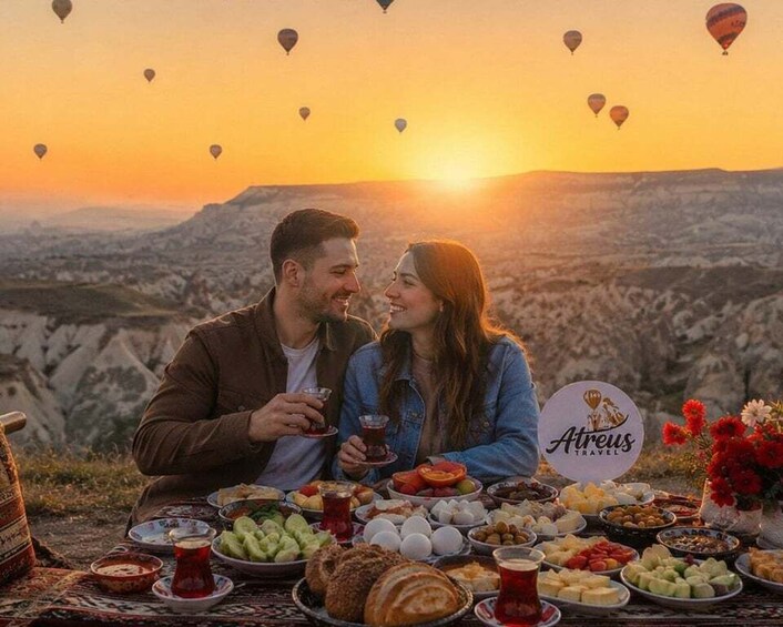 A couple dining outdoors with a view of a mountainous landscape and hot air balloons in the sky. 2