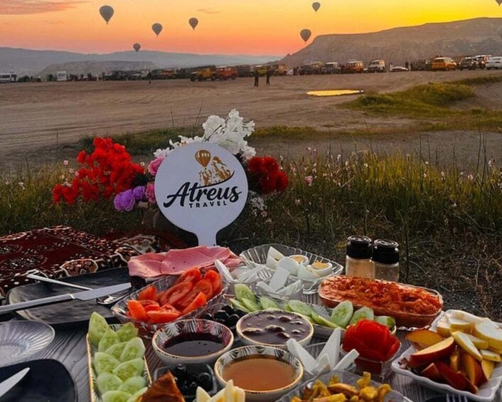 A picnic setup with food and a sign reading 'Ateus Travel' in a field with hot air balloons in the background. 1