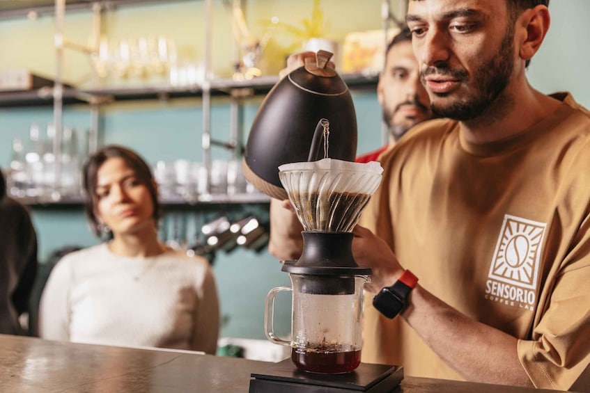 A person in a yellow shirt with a logo, preparing a beverage with a coffee grinder and a pour-over setup. 5