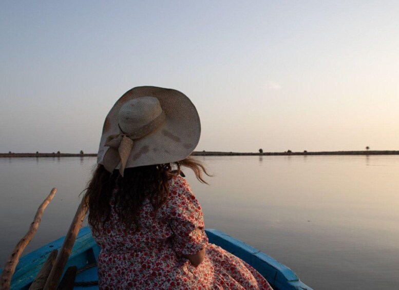 A person in a boat wearing a floral dress and a wide-brimmed hat, with calm water and a distant shoreline. 1