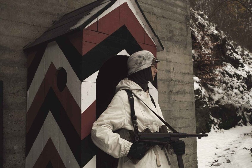 A person in a white jacket with a rifle, standing in front of a building with a chevron pattern. 3