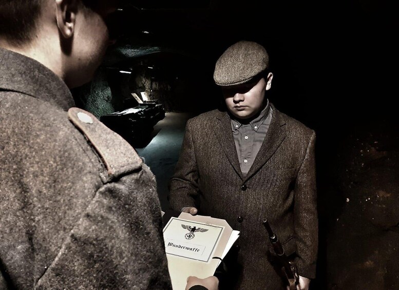Two individuals in a dimly lit setting, one holding a document with a logo. 1