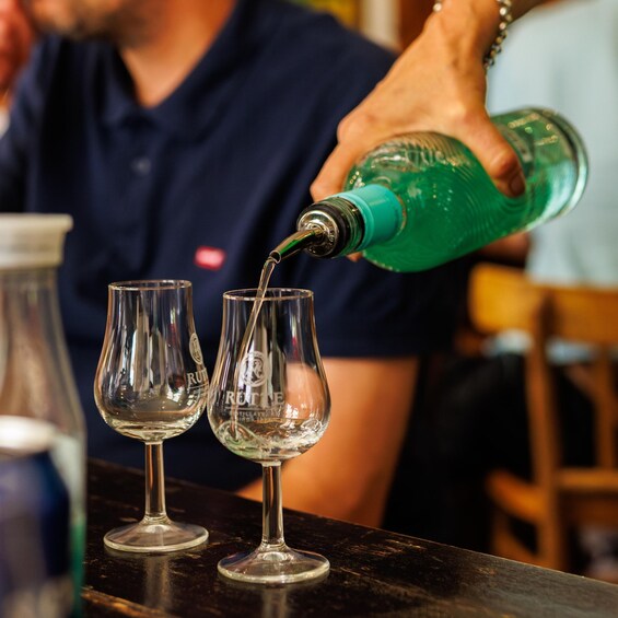 A person pouring a green liquid into two wine glasses on a bar counter. 1